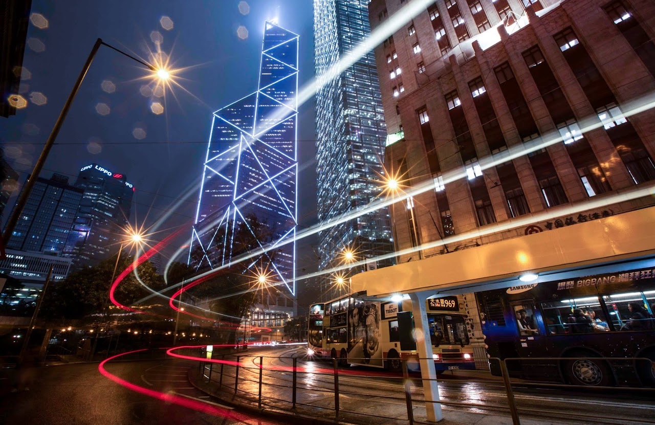 Hong Kong skyscrapers and buses with sweeping light trails at night.