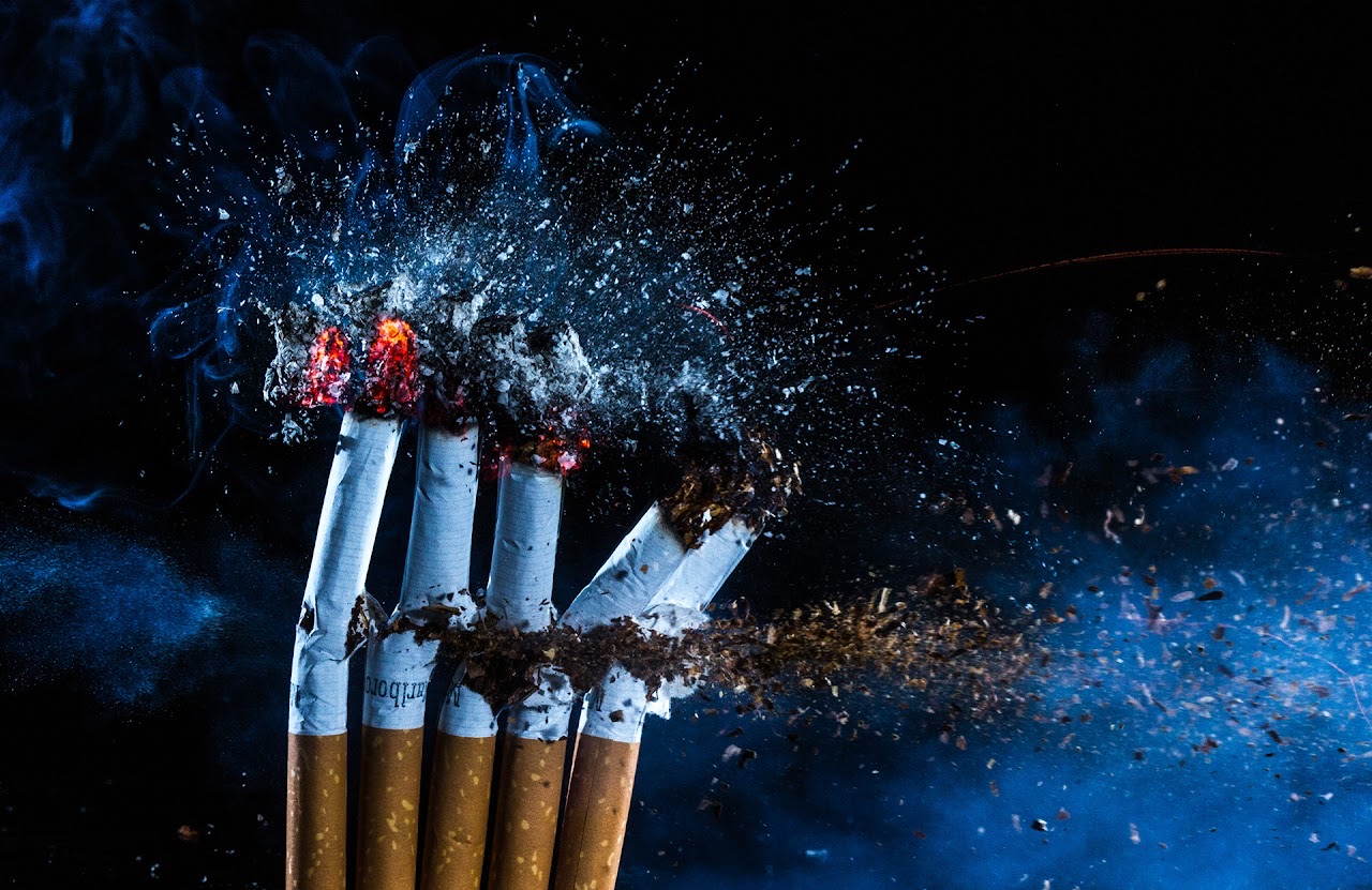 Cigarettes exploding into ash and blue smoke against a dark background.