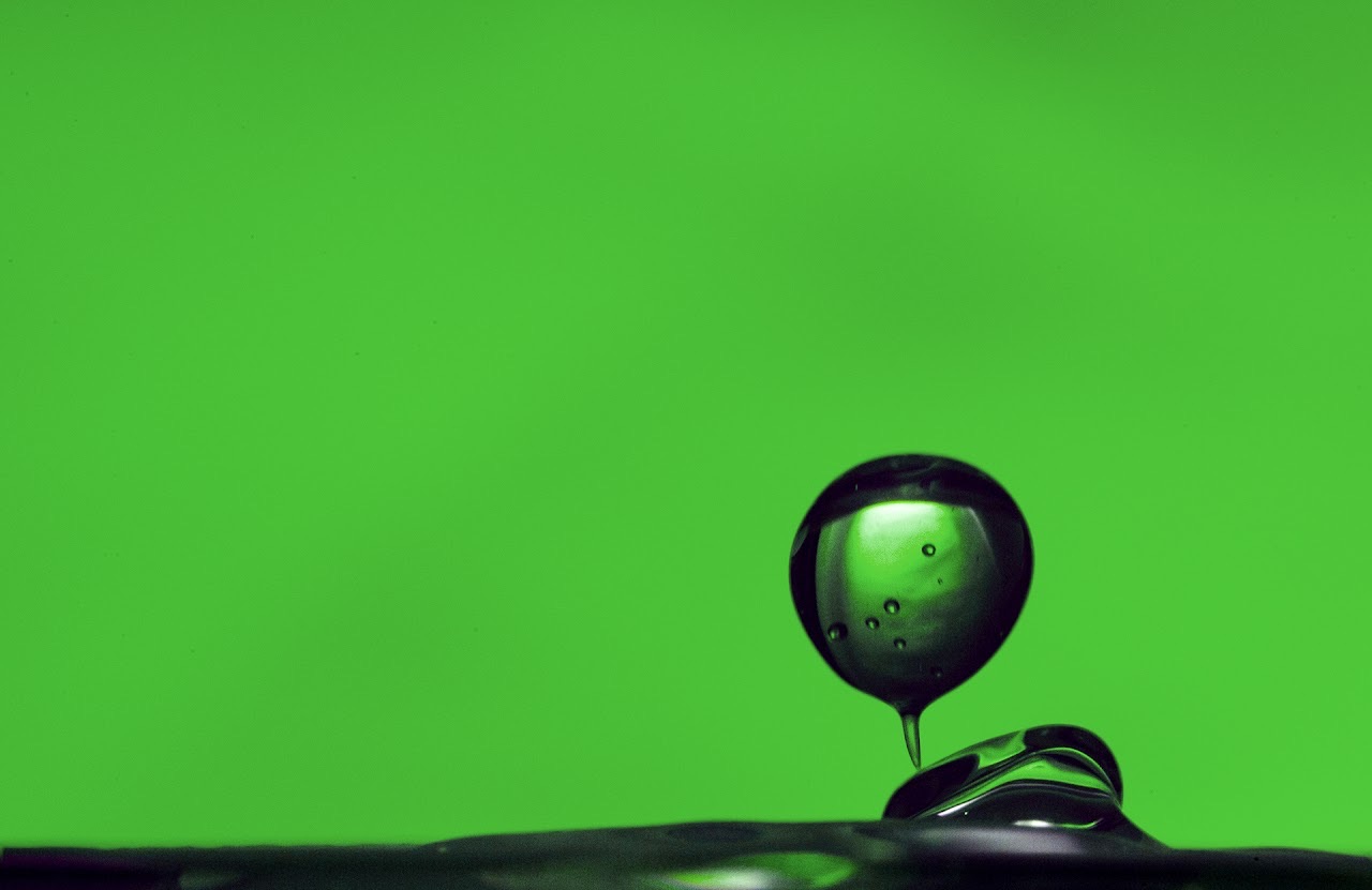 A dark green water droplet hovering over a liquid surface.