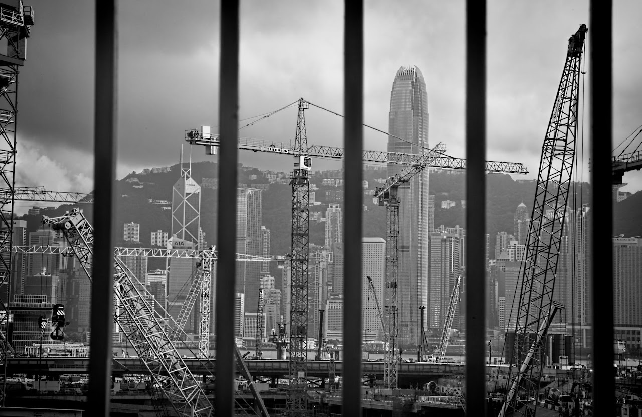 Monochrome Hong Kong skyline with cranes behind vertical bars.