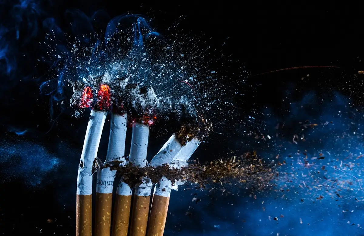 Cigarettes exploding into ash and blue smoke against a dark background.