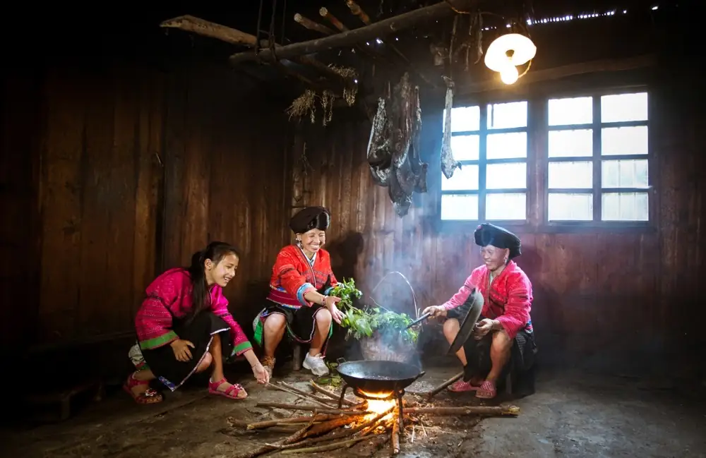 Three women cooking around a fire inside a dark wooden room.