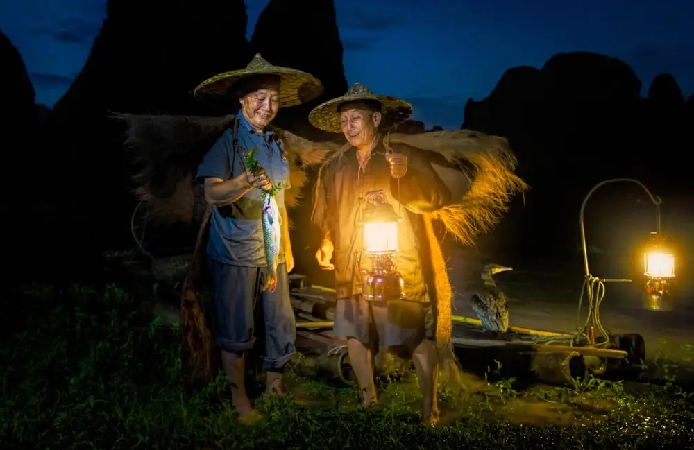 Two fishermen at night with glowing lamps and dark mountains behind them.