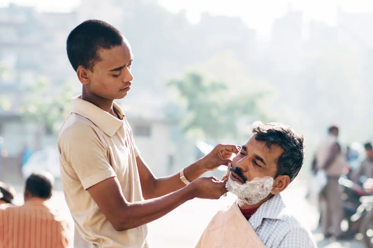 A boy shaving a seated man outdoors.