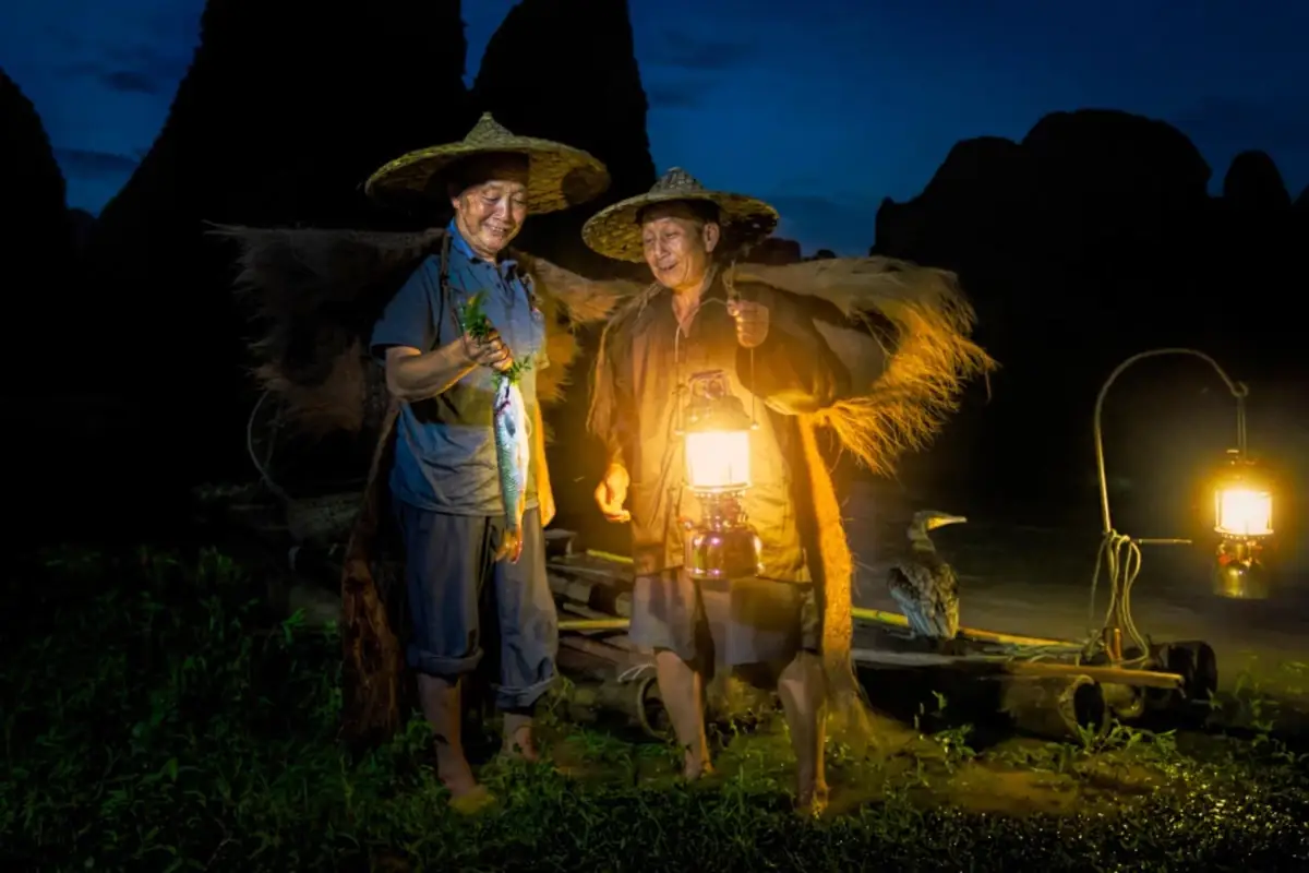 Two fishermen at night holding lanterns beside water.