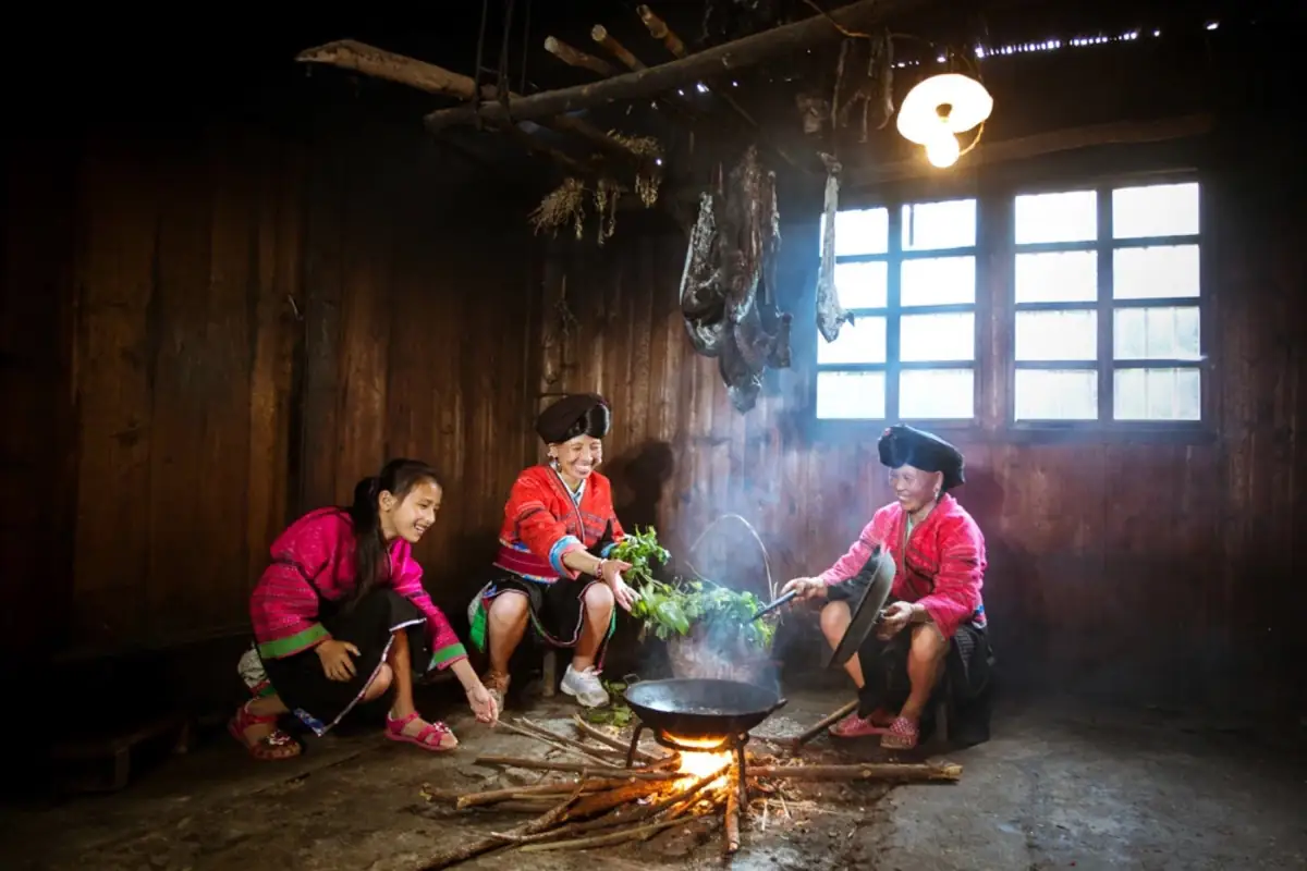 Women cooking around a hearth in a dark wooden room.